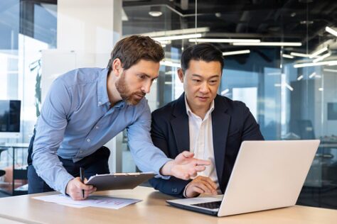Two business professionals collaborating at a laptop in a modern office. One stands beside the desk holding a clipboard and gesturing toward the screen, while the other operates the laptop. The image illustrates teamwork, workflow evaluation, and process improvement.